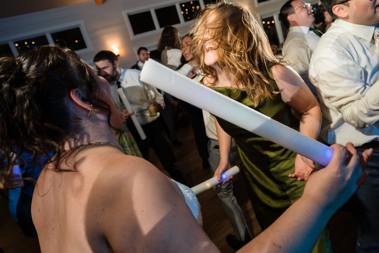 A bride and a guest dance together holding glowing foam light sticks at a wedding reception.