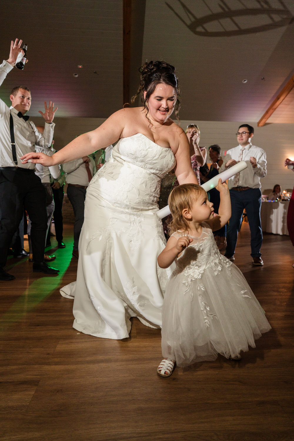 A bride in a white wedding gown dances on a wooden floor with a young child in a matching white dress who is holding a glow stick.