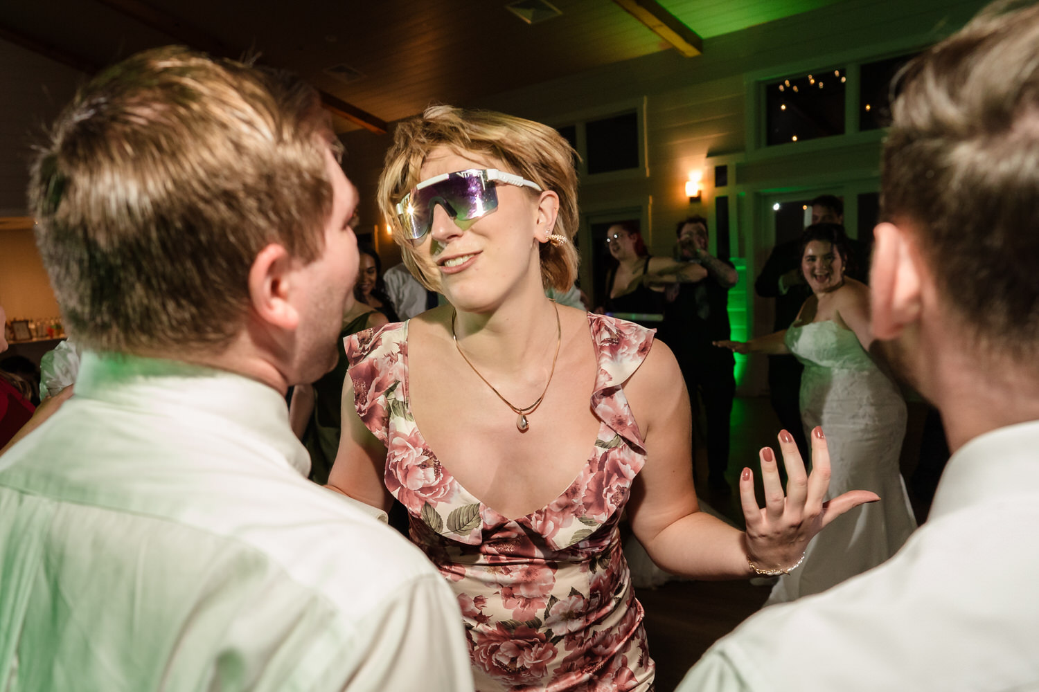 A woman in a floral dress and large reflective sunglasses dances between two men at a wedding reception.