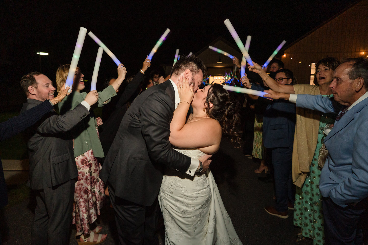 A bride and groom kissing surrounded by wedding guests holding glowing foam sticks.