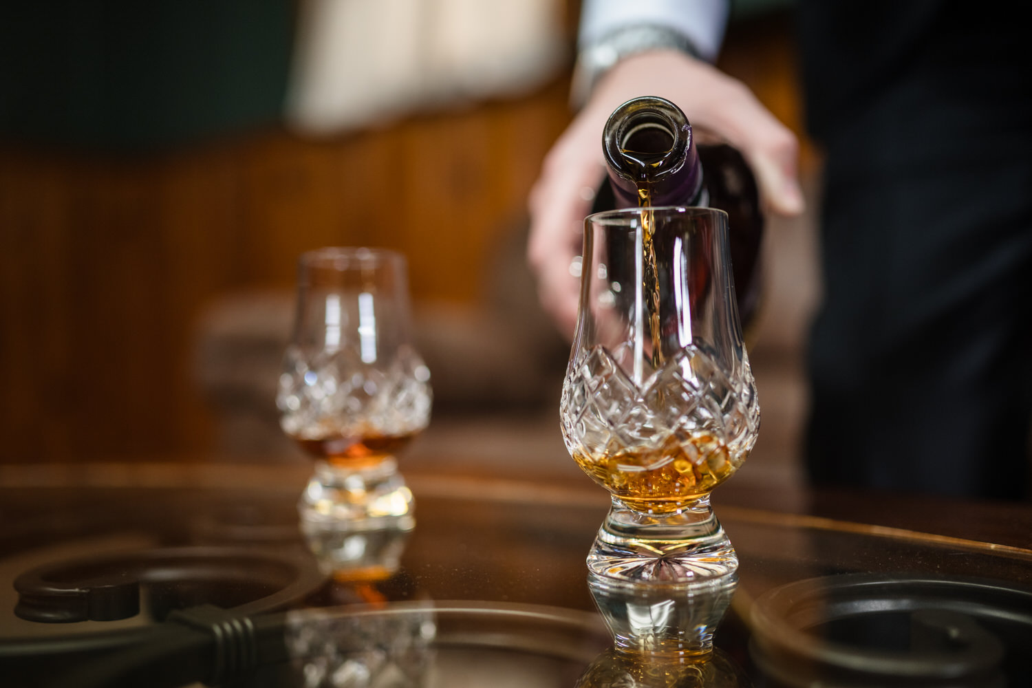 A person pours amber liquid from a bottle into a crystal glass on a table, with another filled glass nearby.
