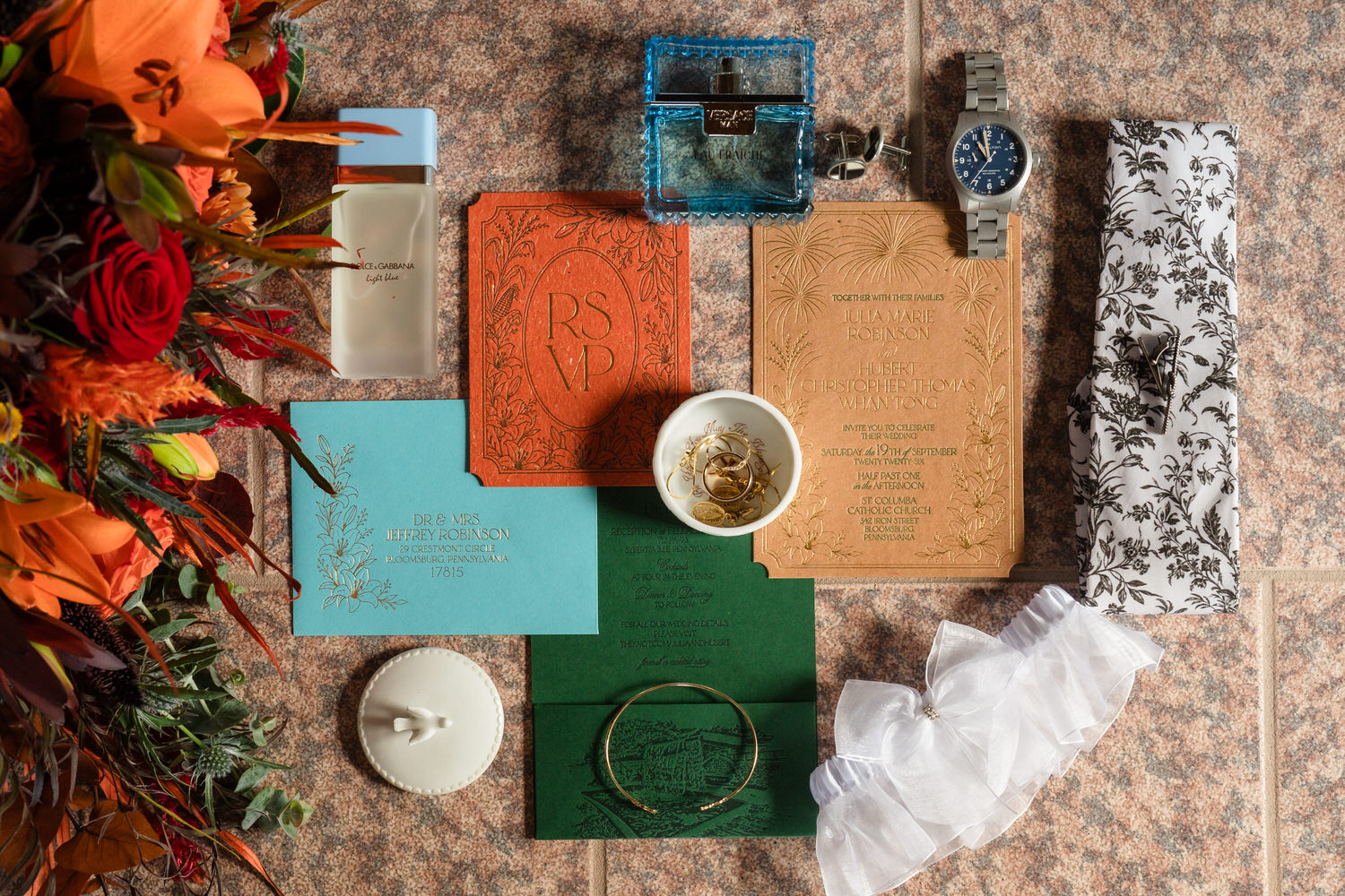 Wedding stationery, perfume bottles, a watch, jewelry, and a floral bouquet arranged on a stone surface.