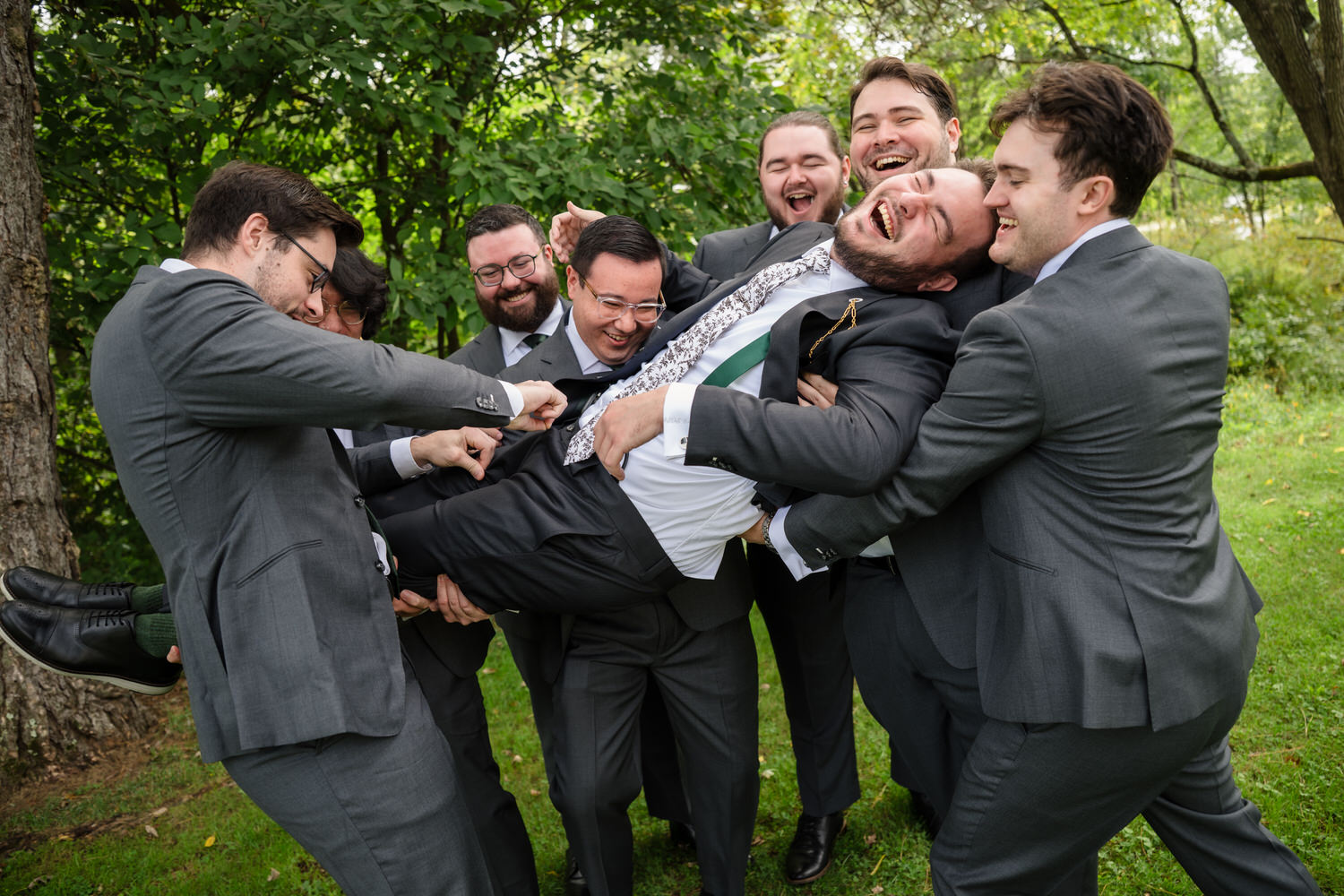 Six groomsmen in grey suits laugh as they playfully lift and carry another groomsman in a suit outdoors.