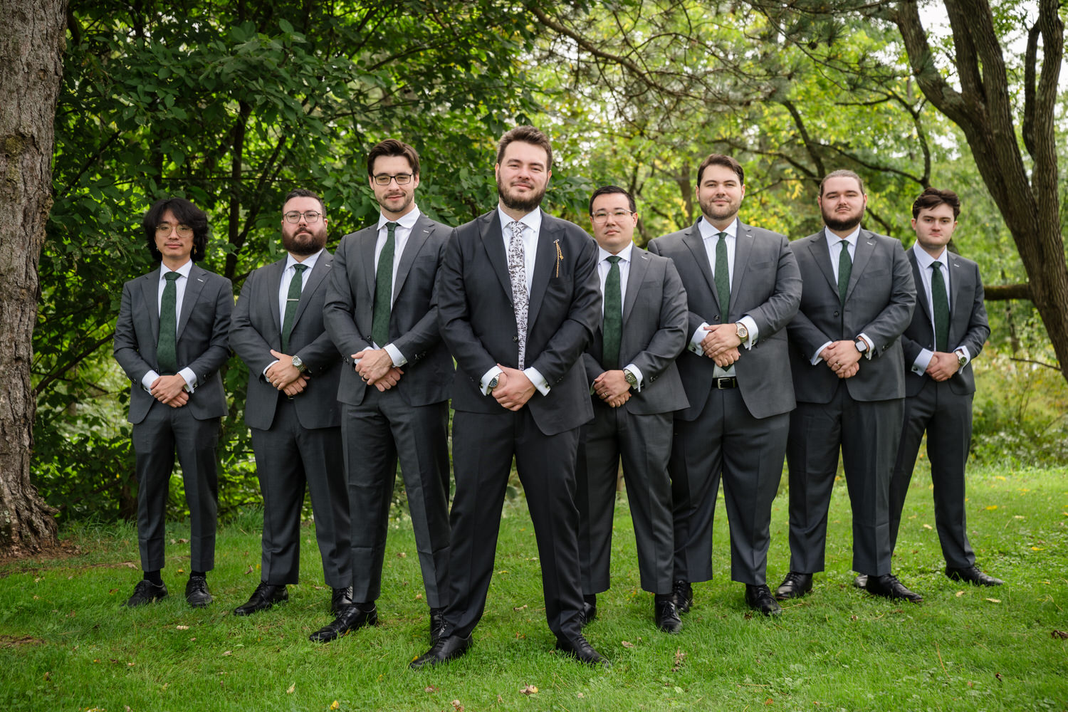 Eight men in matching grey suits and green ties stand in a line on a grassy lawn.