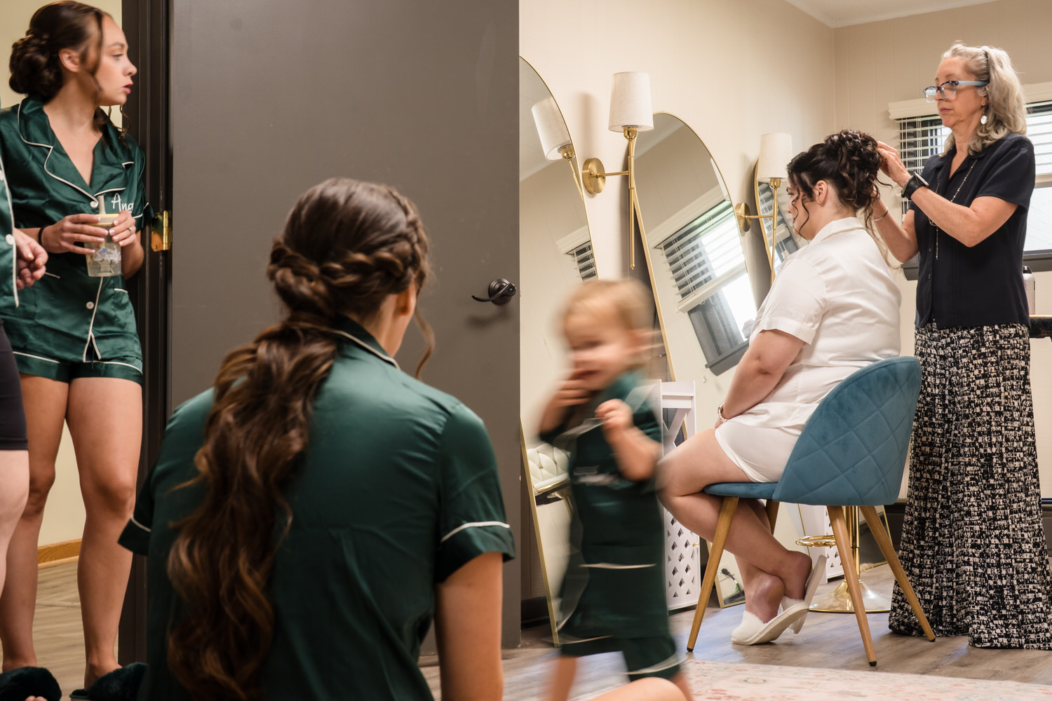 A stylist styles the hair of a bride in a white robe, while two bridesmaids in green pajamas watch and a child runs past in the foreground.