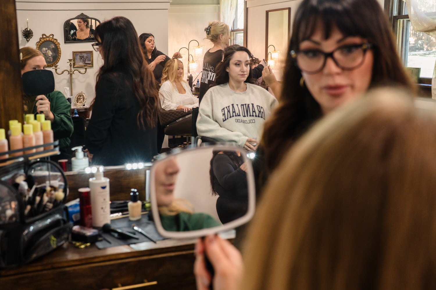 A hairstylist works on a clients hair in a salon while others wait, as seen through a reflection in a handheld mirror held by a woman in the foreground.