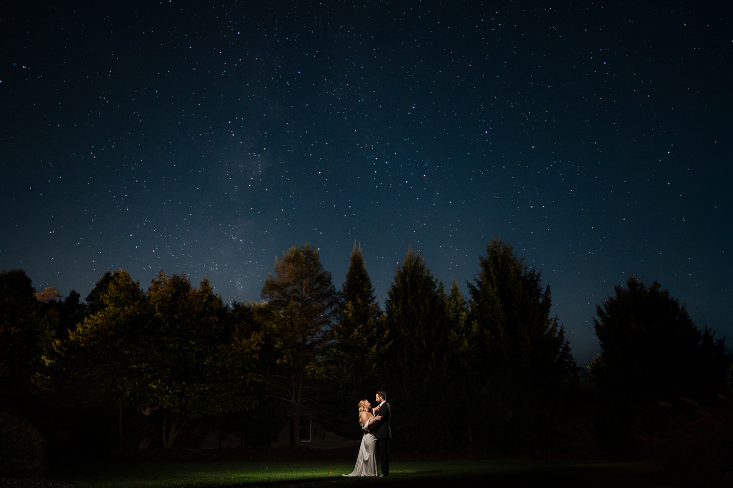A couple embraces in a spotlight on a lawn under a vast, star-filled night sky.