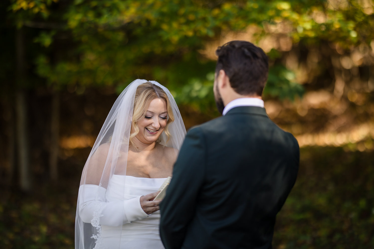 A bride in a white gown and veil smiles while reading from a small booklet to her partner during an outdoor wedding ceremony.