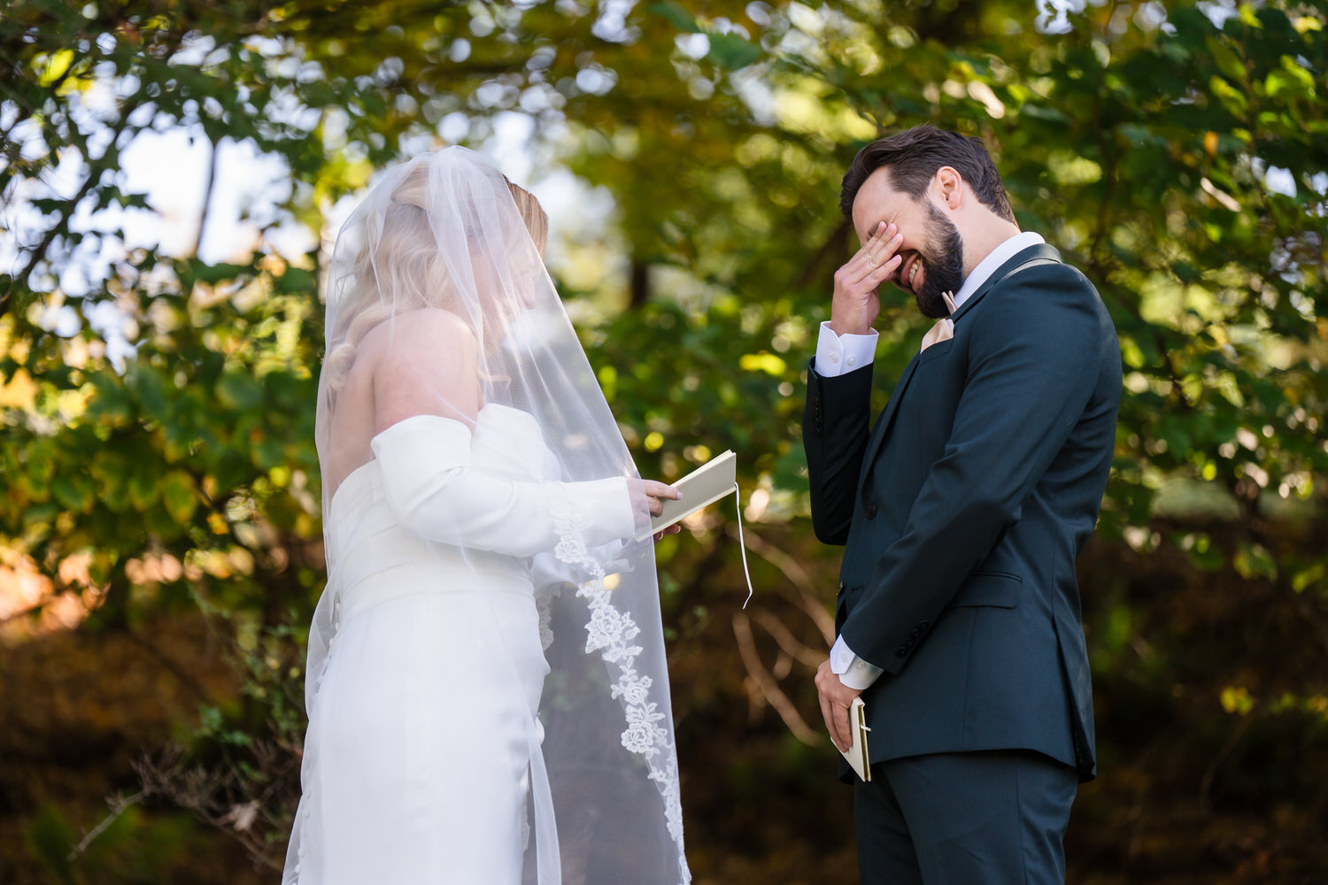 A bride reading vows to a groom who is covering his face with his hand while laughing or crying during an outdoor wedding ceremony.