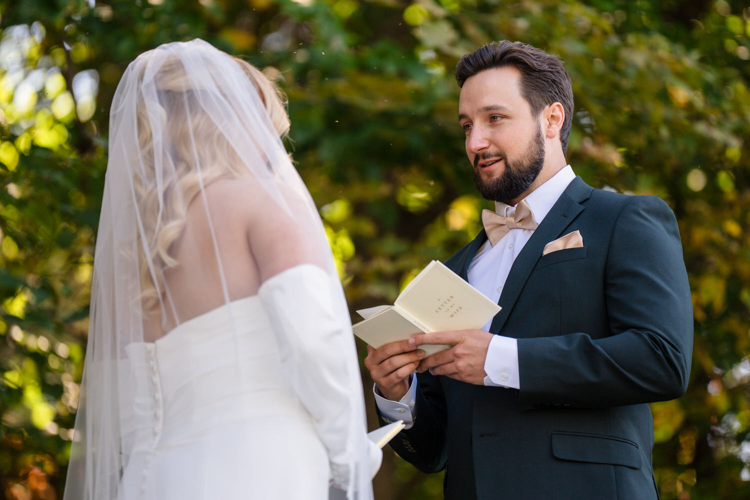 A groom in a tuxedo reads from a small book labeled Letter to my Wife while facing his bride during an outdoor wedding ceremony.