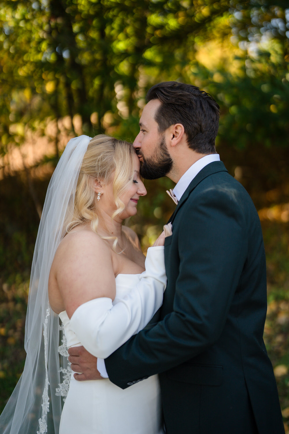A groom kisses the forehead of his bride as they embrace outdoors.