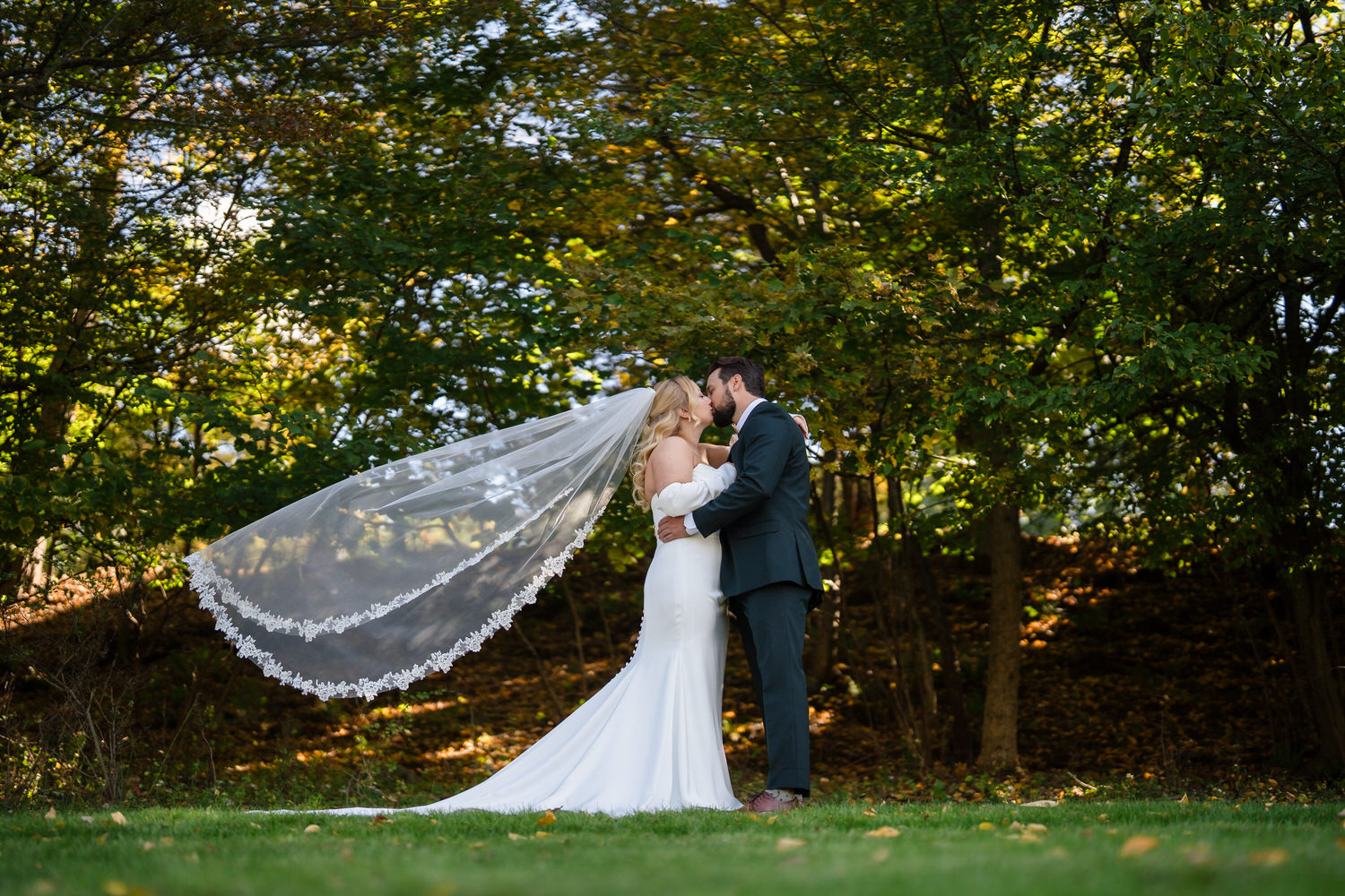 A bride in a white wedding gown and a groom in a dark suit kissing outdoors, with the brides long lace-edged veil trailing behind her.