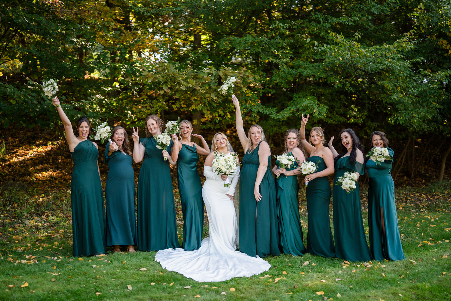 A bride in a white wedding gown and nine bridesmaids in long, dark green dresses pose together in a wooded area.