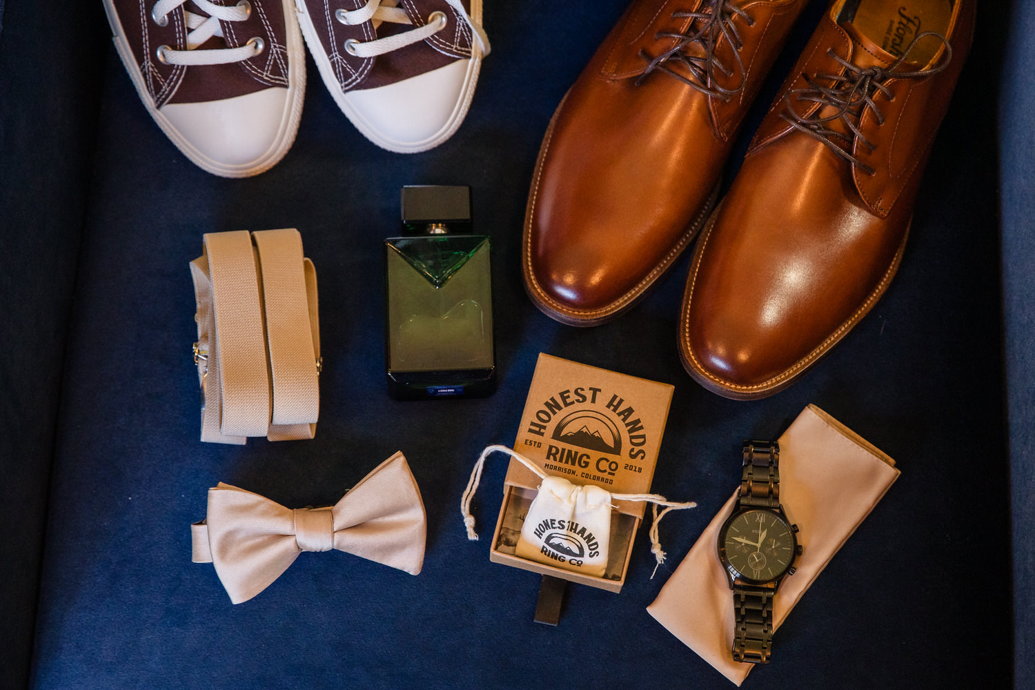 Mens wedding accessories including brown dress shoes, white sneakers, a bow tie, suspenders, a cologne bottle, a watch, and a ring box from Honest Hands Ring Co.