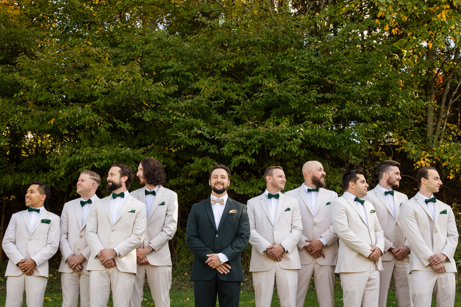 A group of nine groomsmen stand in a line outdoors in front of a tree, with the central man wearing a dark green suit and the others wearing light beige suits with green bow ties.