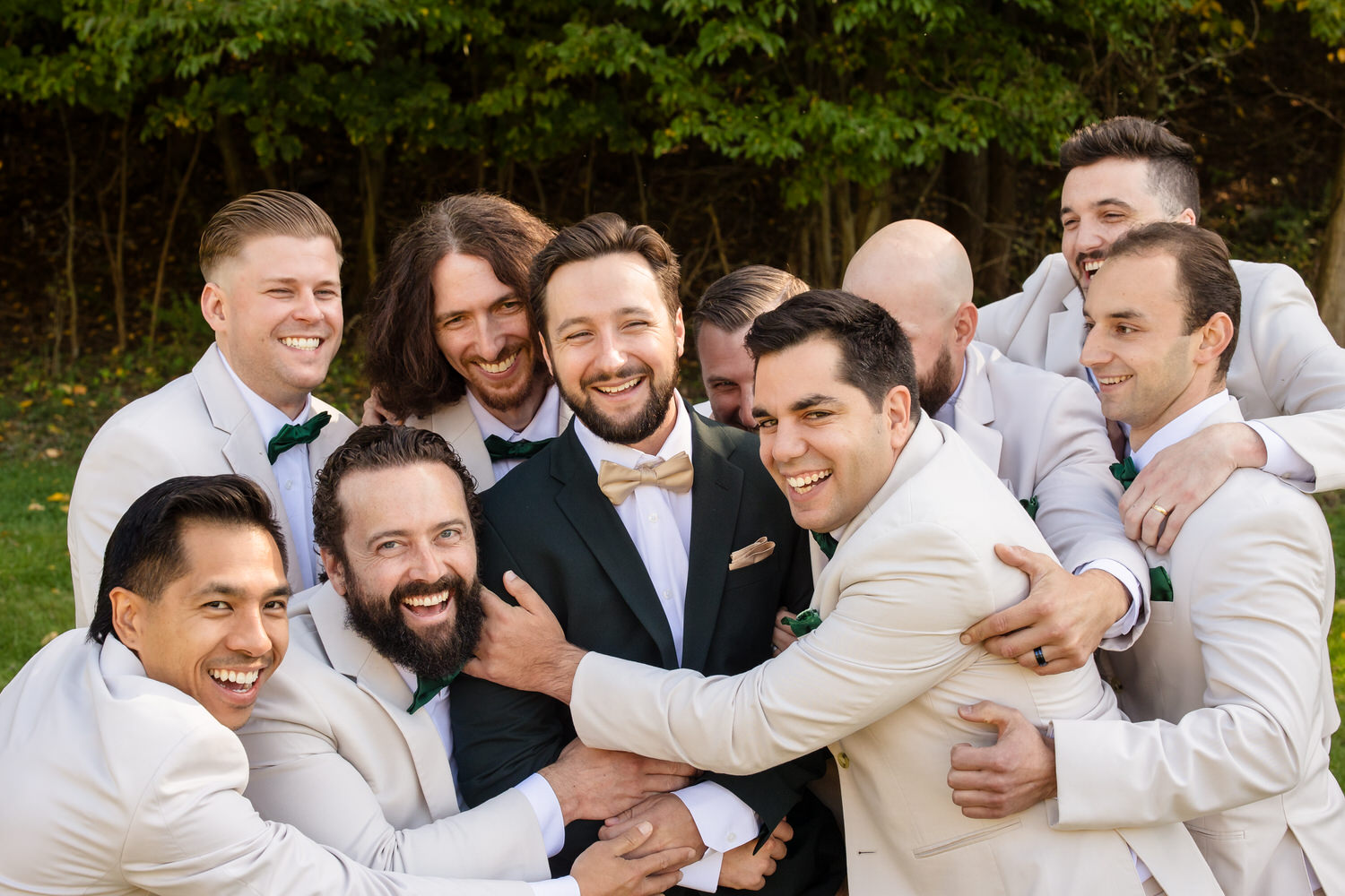 A groom in a dark suit surrounded by his groomsmen in light-colored suits, all smiling and embracing him.