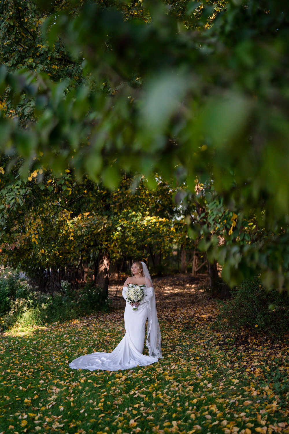 A bride in a white wedding gown and veil holding a floral bouquet, standing on a path covered in autumn leaves framed by trees.