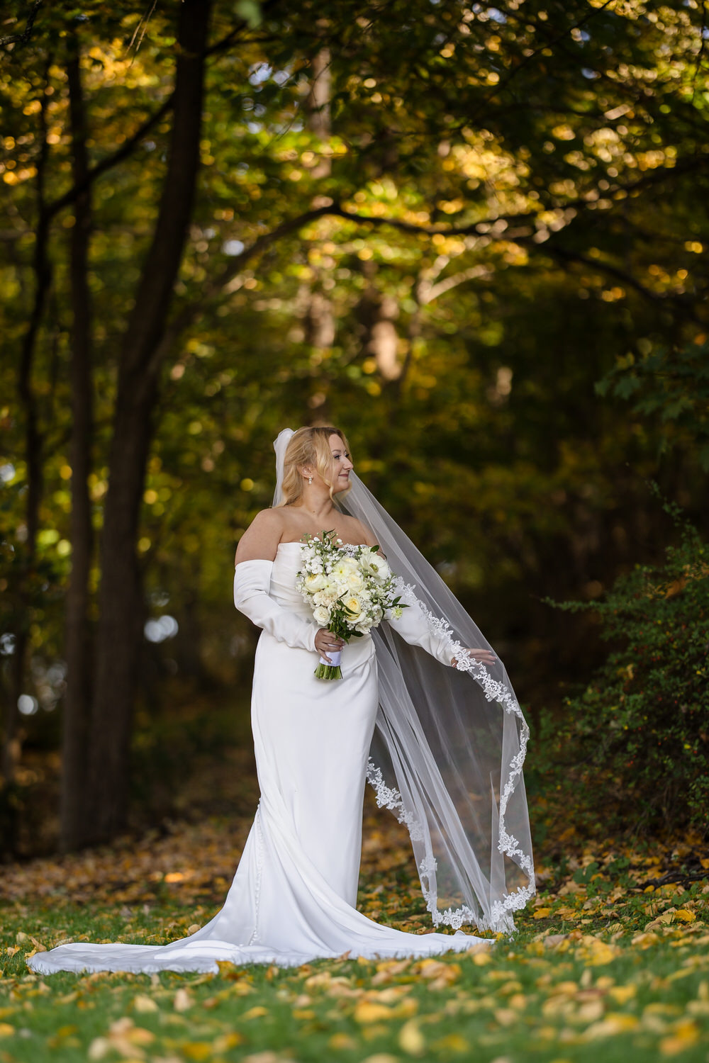 A bride in a white wedding gown and veil holding a bouquet of white flowers in an autumn forest.