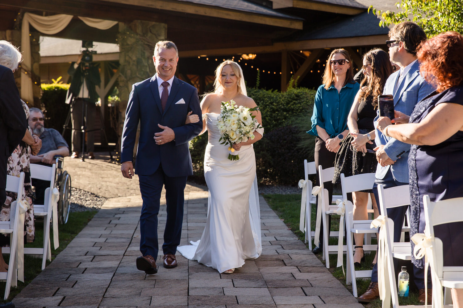 A bride in a white gown and veil walks down an outdoor stone aisle on the arm of a man in a navy suit, with wedding guests watching from chairs on either side.