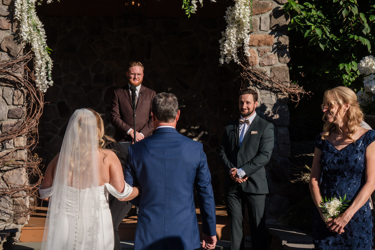 A bride and a man in a blue suit walk toward an officiant and a groom standing before a stone archway, with a guest standing to the right.