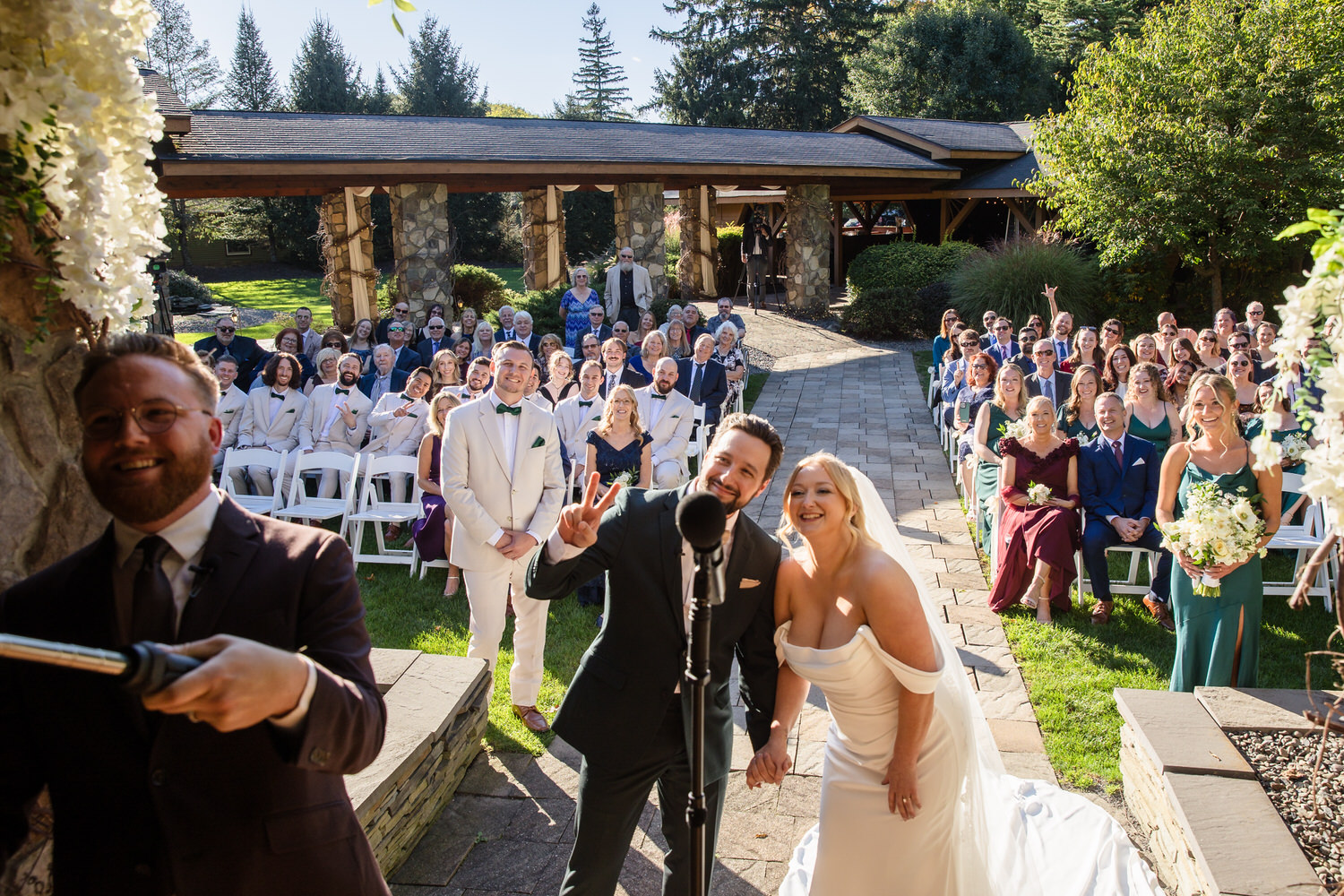 A groom making a peace sign and a bride in a white wedding dress stand at an altar while a guest records the ceremony with a phone, with seated wedding guests in the background.