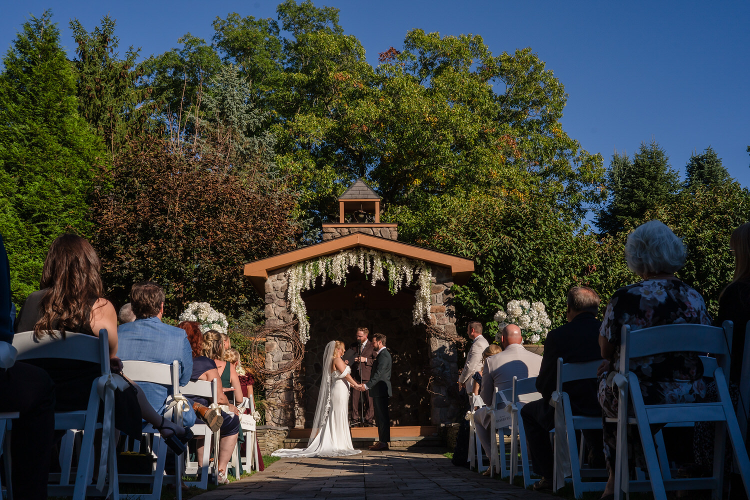 A bride and groom exchange vows under a stone garden structure while guests seated in rows of white chairs watch from behind.