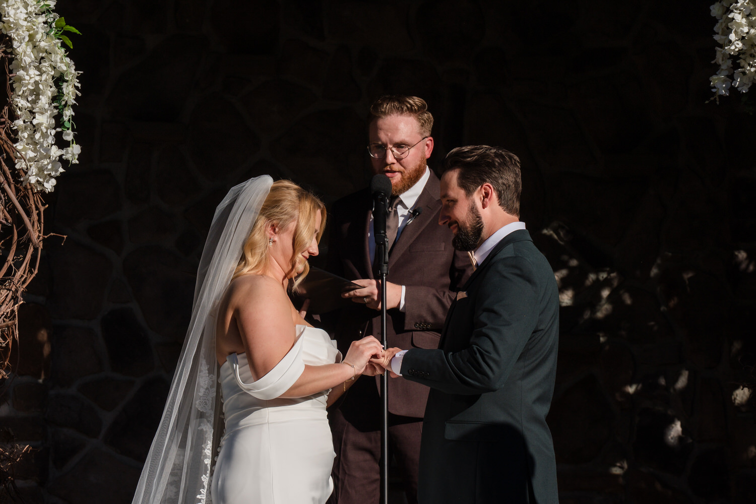 A bride and groom exchange wedding rings at the altar while an officiant stands behind them.