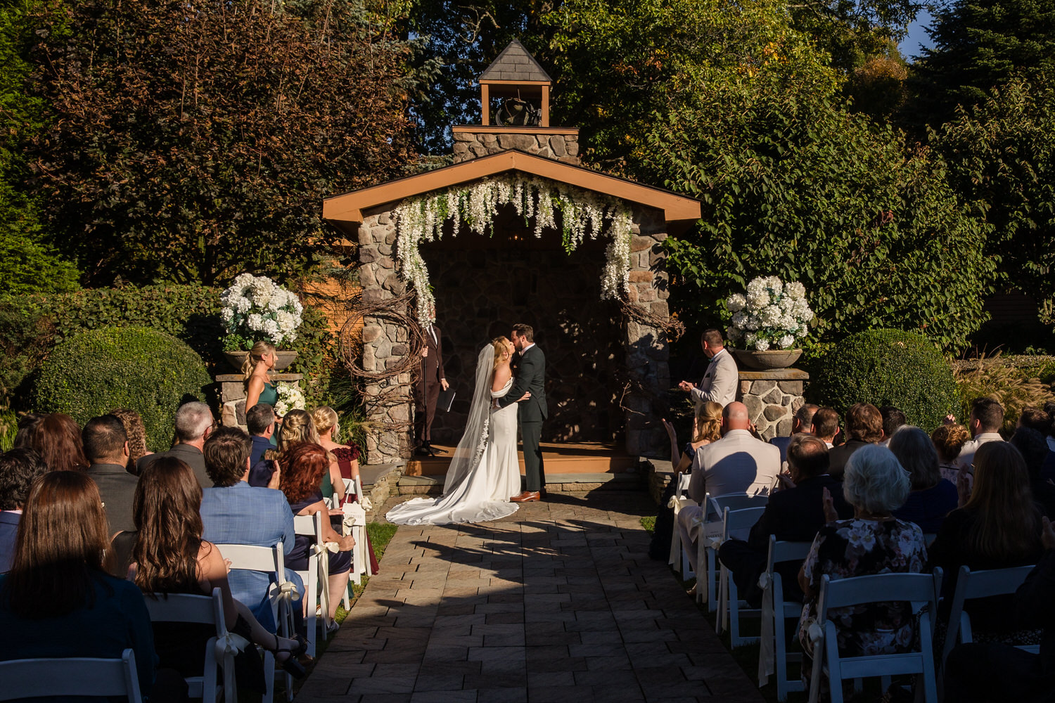 A bride and groom kiss under a stone arbor decorated with white flowers, while guests seated on both sides of a stone aisle look on during an outdoor wedding ceremony.