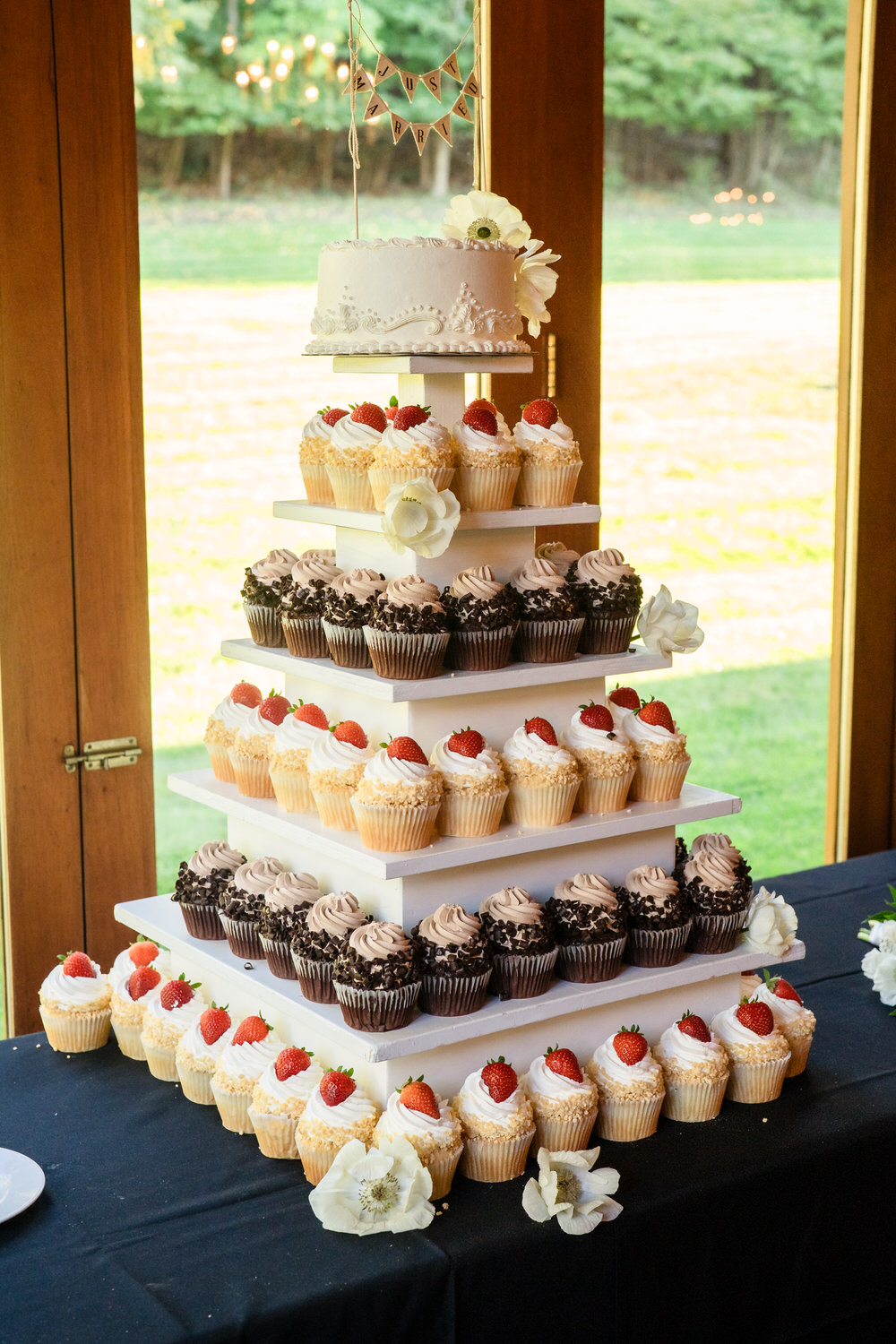 A five-tiered stand holding a small white wedding cake on top and rows of frosted cupcakes topped with strawberries on the shelves below.