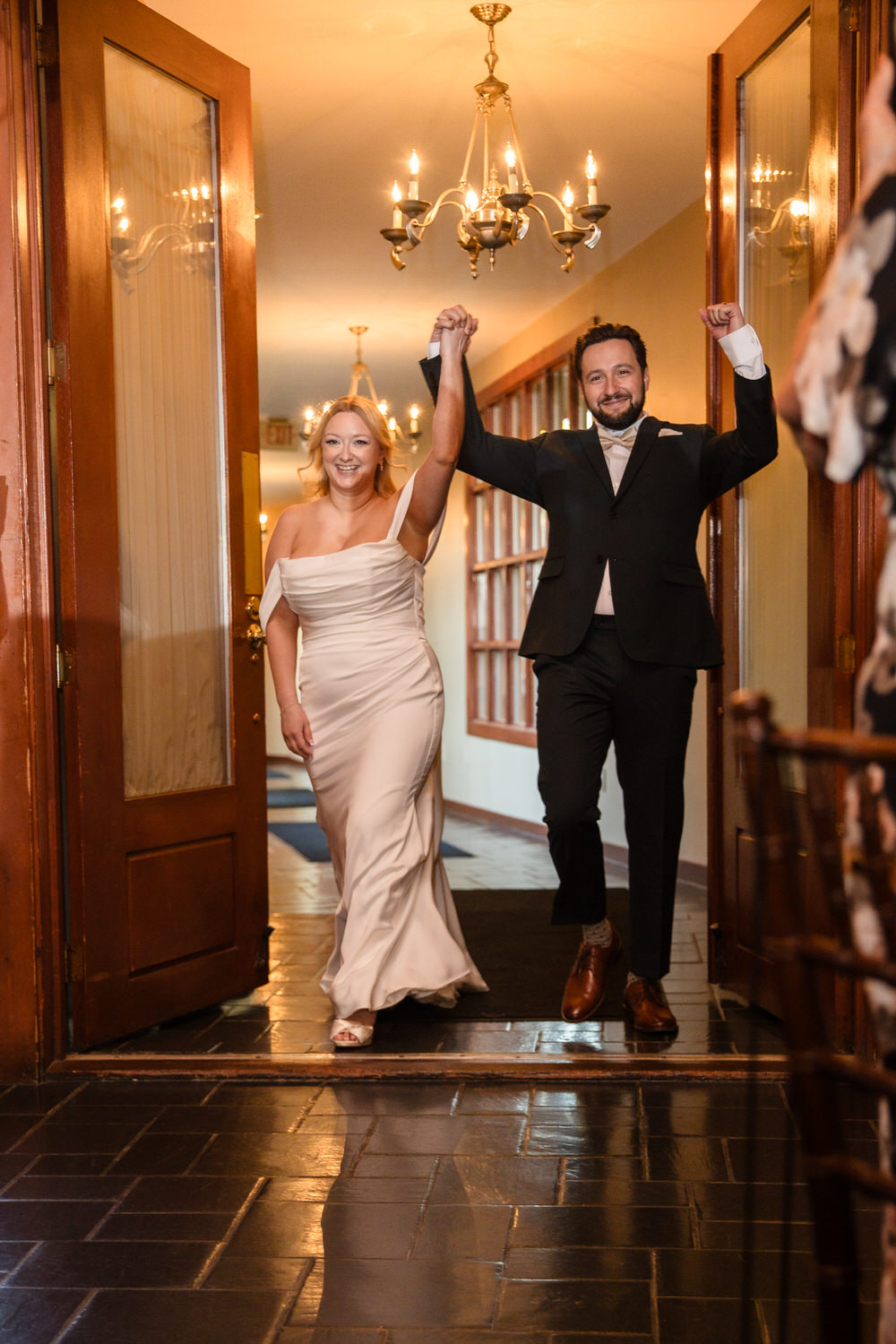 A bride and groom walk through open double doors into a room, holding their joined hands high in celebration.