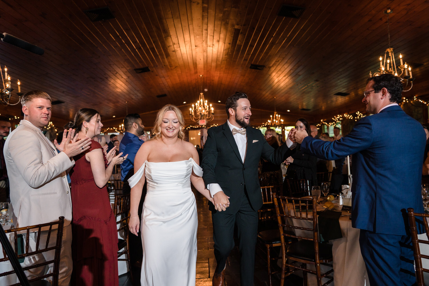 A bride and groom walk through a reception hall, holding hands and smiling, as guests clap for them.