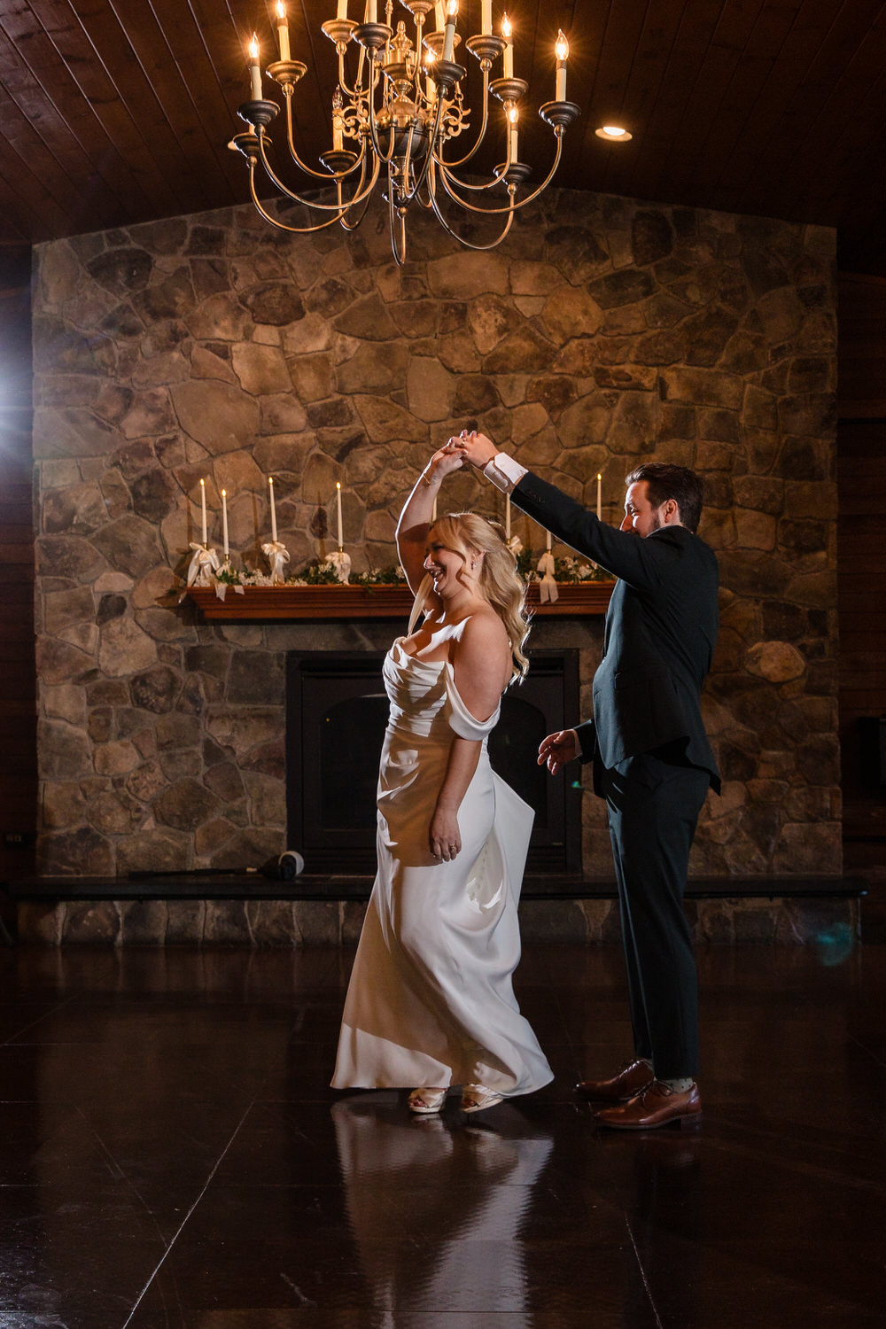 A bride in a white wedding gown and a groom in a black suit dancing in front of a stone fireplace.