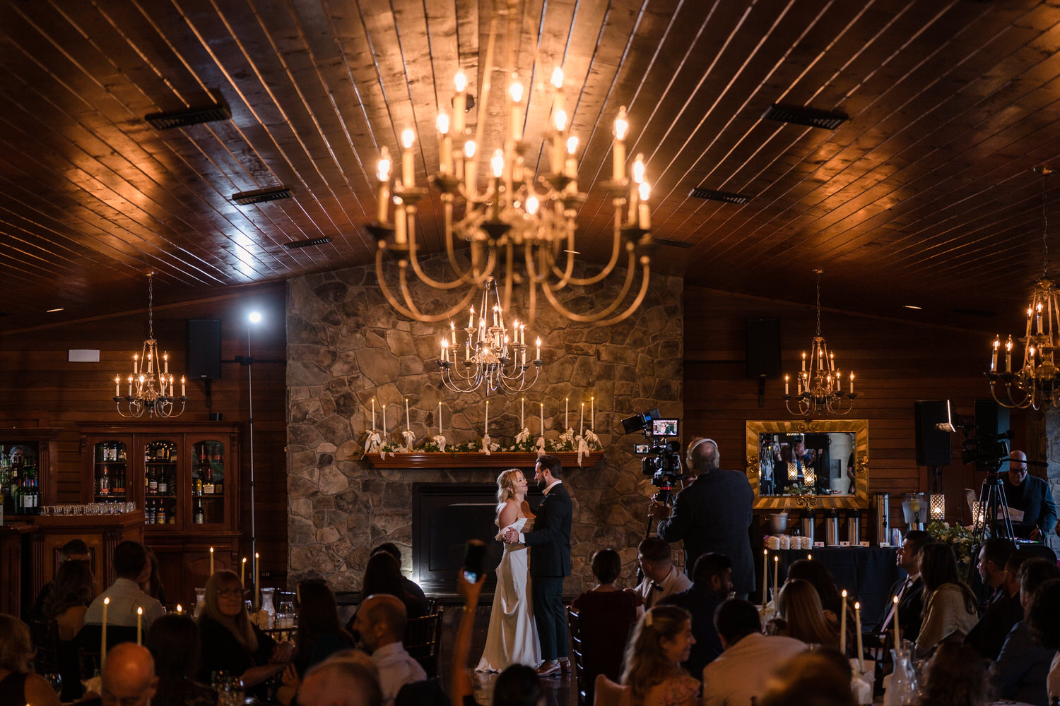 A bride and groom share their first dance in a wood-paneled room with stone fireplaces and a videographer filming nearby.