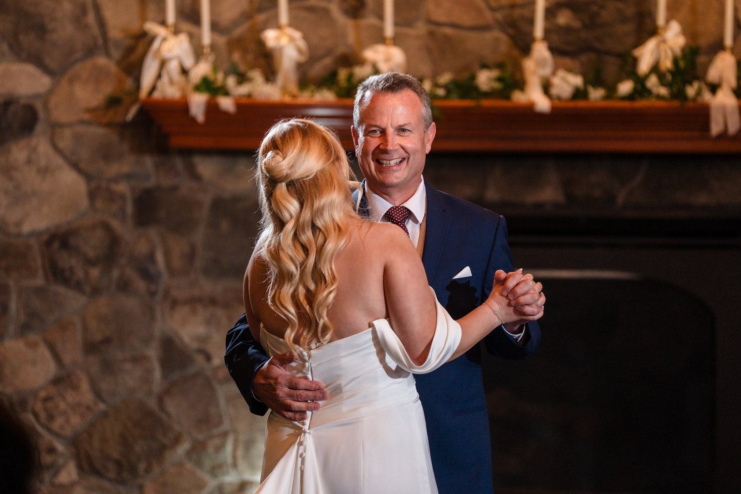 A bride and her father smiling while dancing together at a wedding reception in front of a stone fireplace.