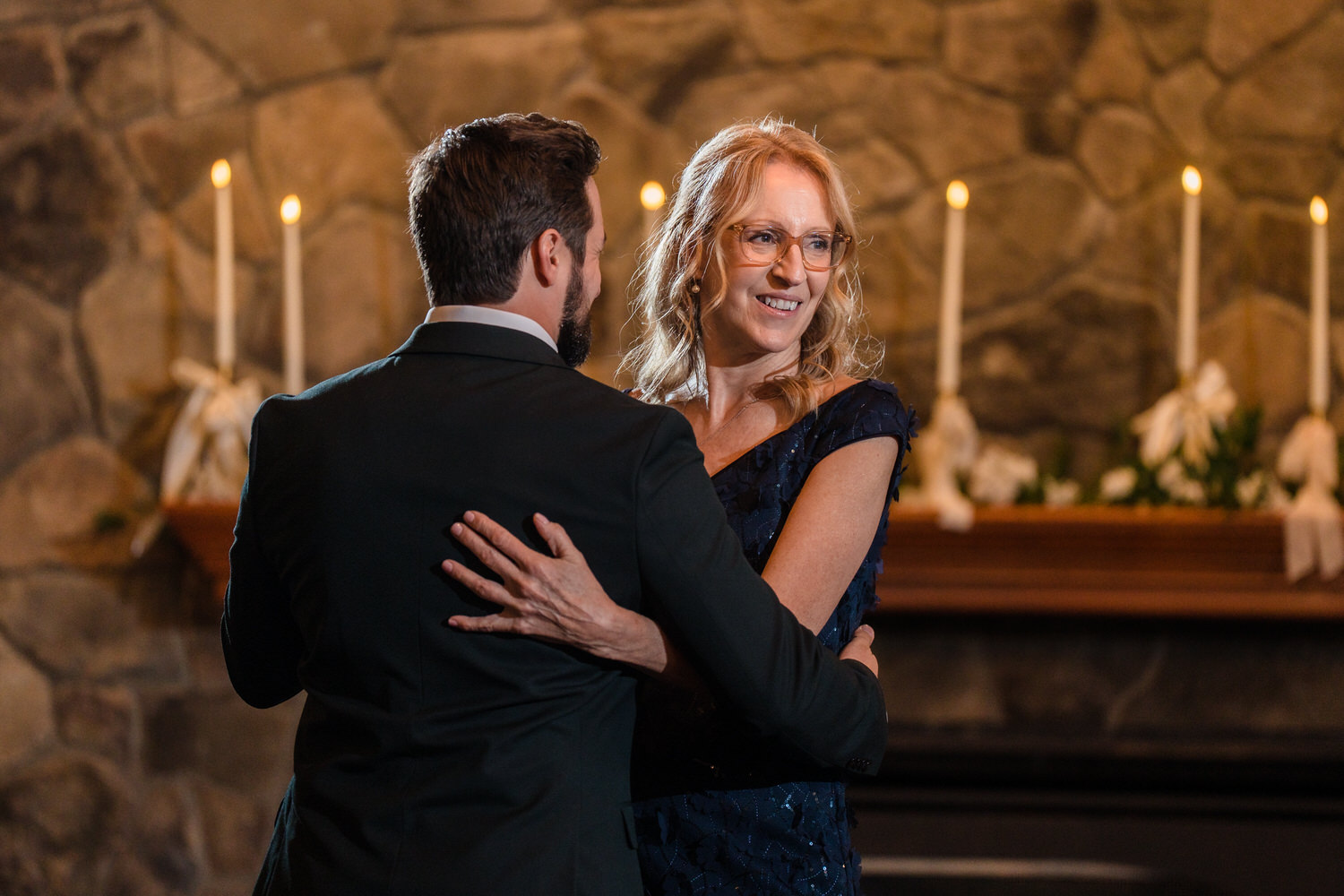 A man in a suit and a woman in a dark dress dance together in front of a stone fireplace with lit candles.