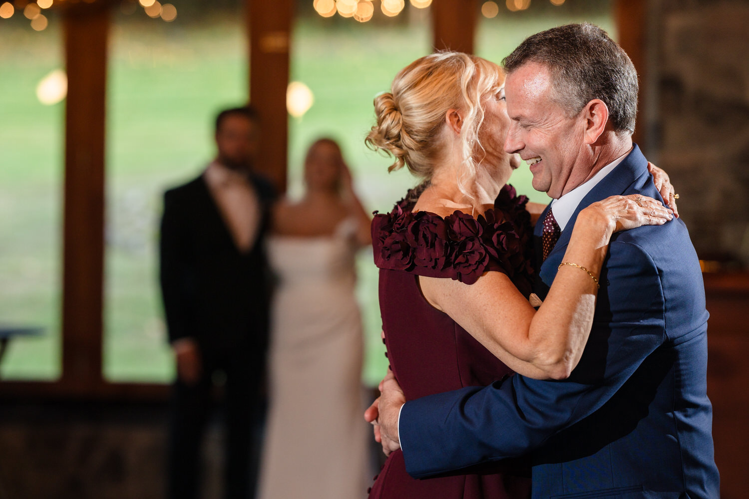 A man in a blue suit and a woman in a maroon dress dance together while smiling, with a bride and groom blurred in the background.