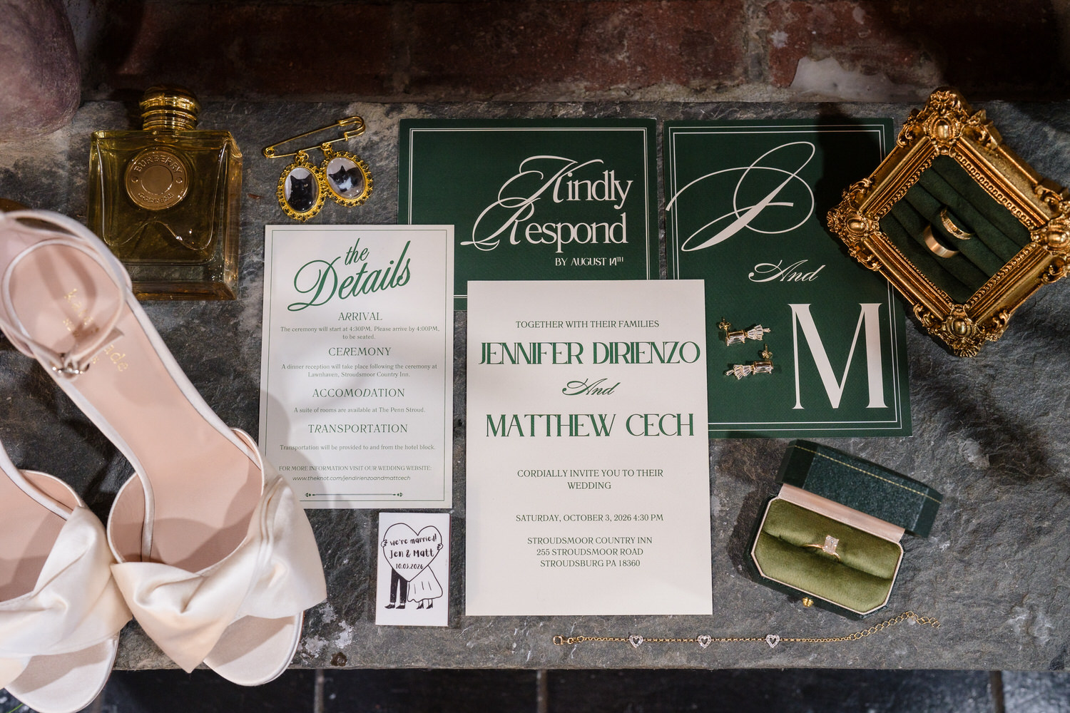 A flat lay of wedding stationery, including an invitation for Jennifer Dirienzo and Matthew Cech, a details card, an RSVP card, a bridal shoe, a ring box with a diamond ring, two wedding bands, a bracelet, and small pins.