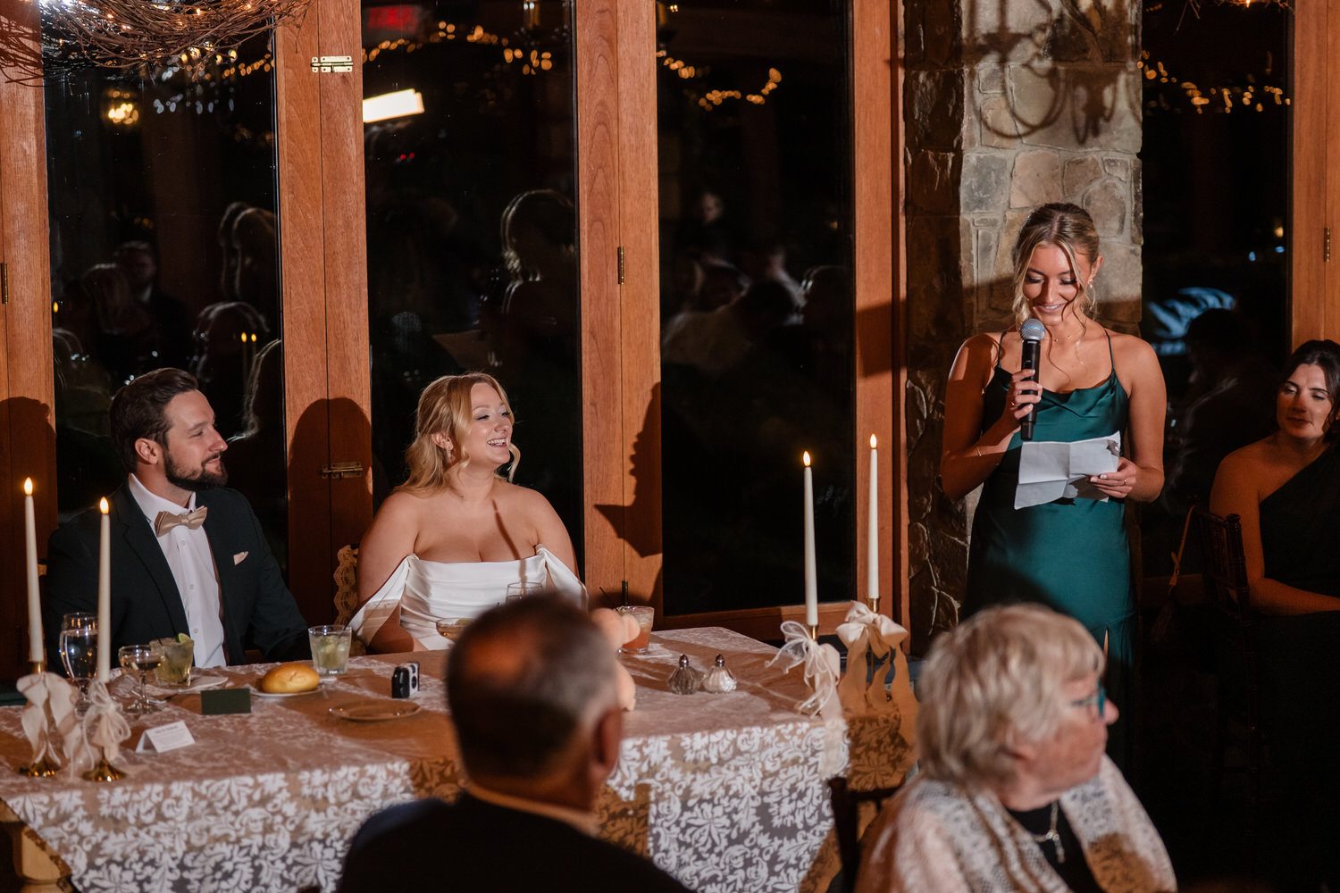 A woman in a green dress stands at a wedding reception giving a speech with a microphone, while a bride and groom sit at a table nearby.
