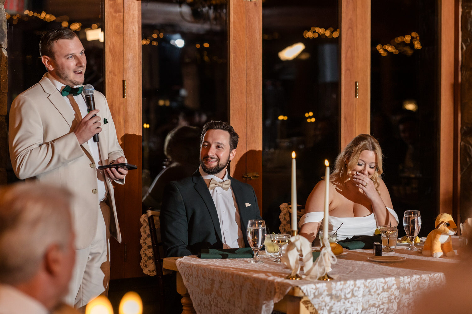 A man in a tan suit stands at a wedding reception giving a speech while a seated couple at a table looks on and smiles.