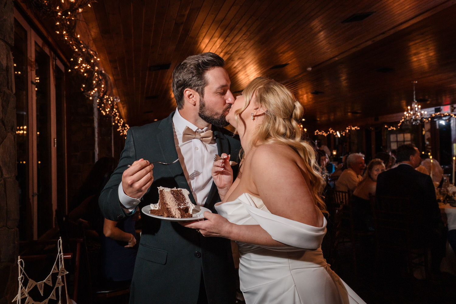 A bride and groom in wedding attire kiss while sharing a piece of chocolate cake at a dimly lit reception.