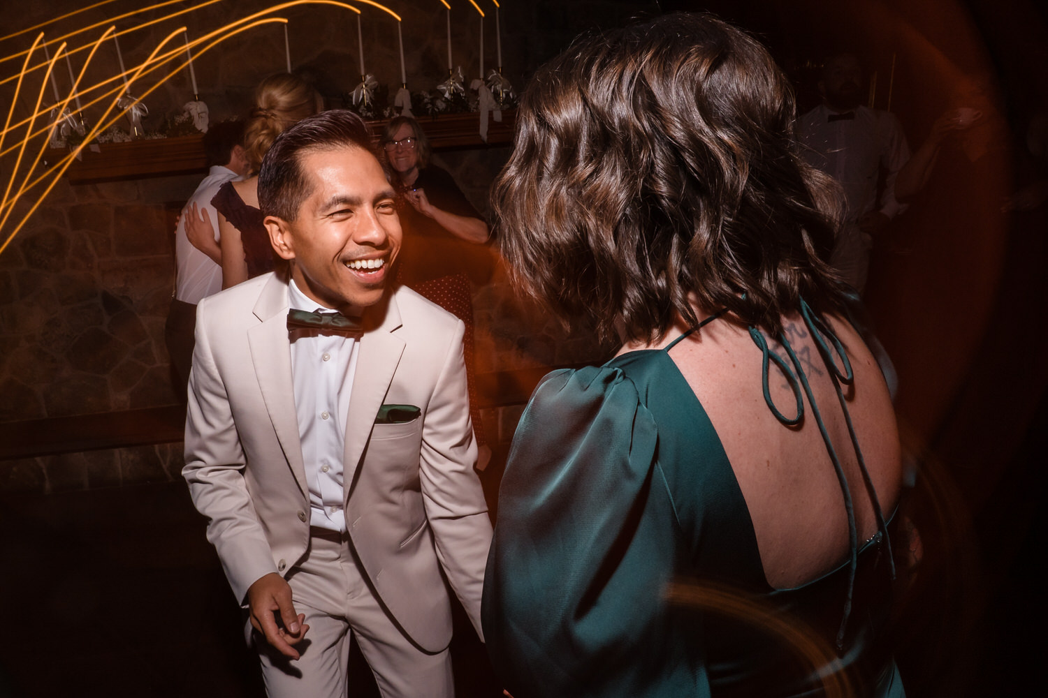 A man in a cream tuxedo and green bowtie smiles at a woman in a green dress while dancing at a party.