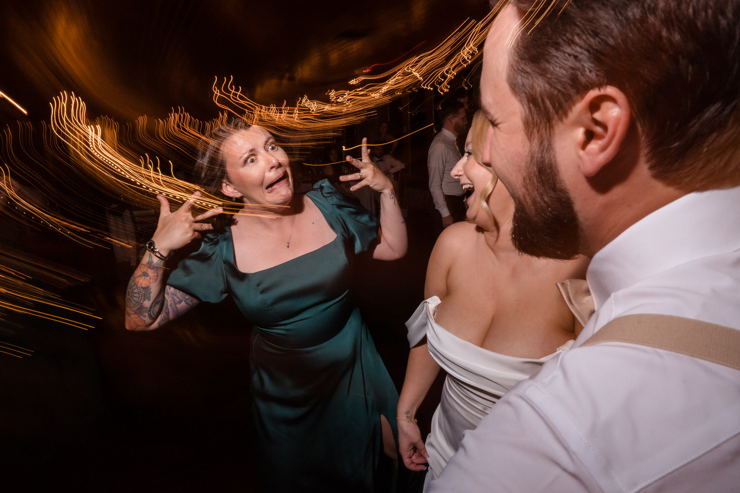 A woman in a green dress makes a playful face while dancing with a bride and groom at a reception.