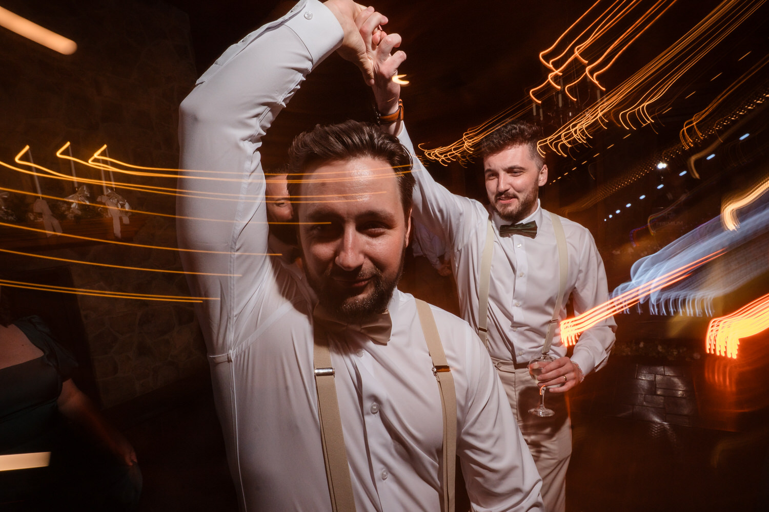 Two men in white dress shirts and suspenders dancing at a party with long-exposure light streaks in the background.