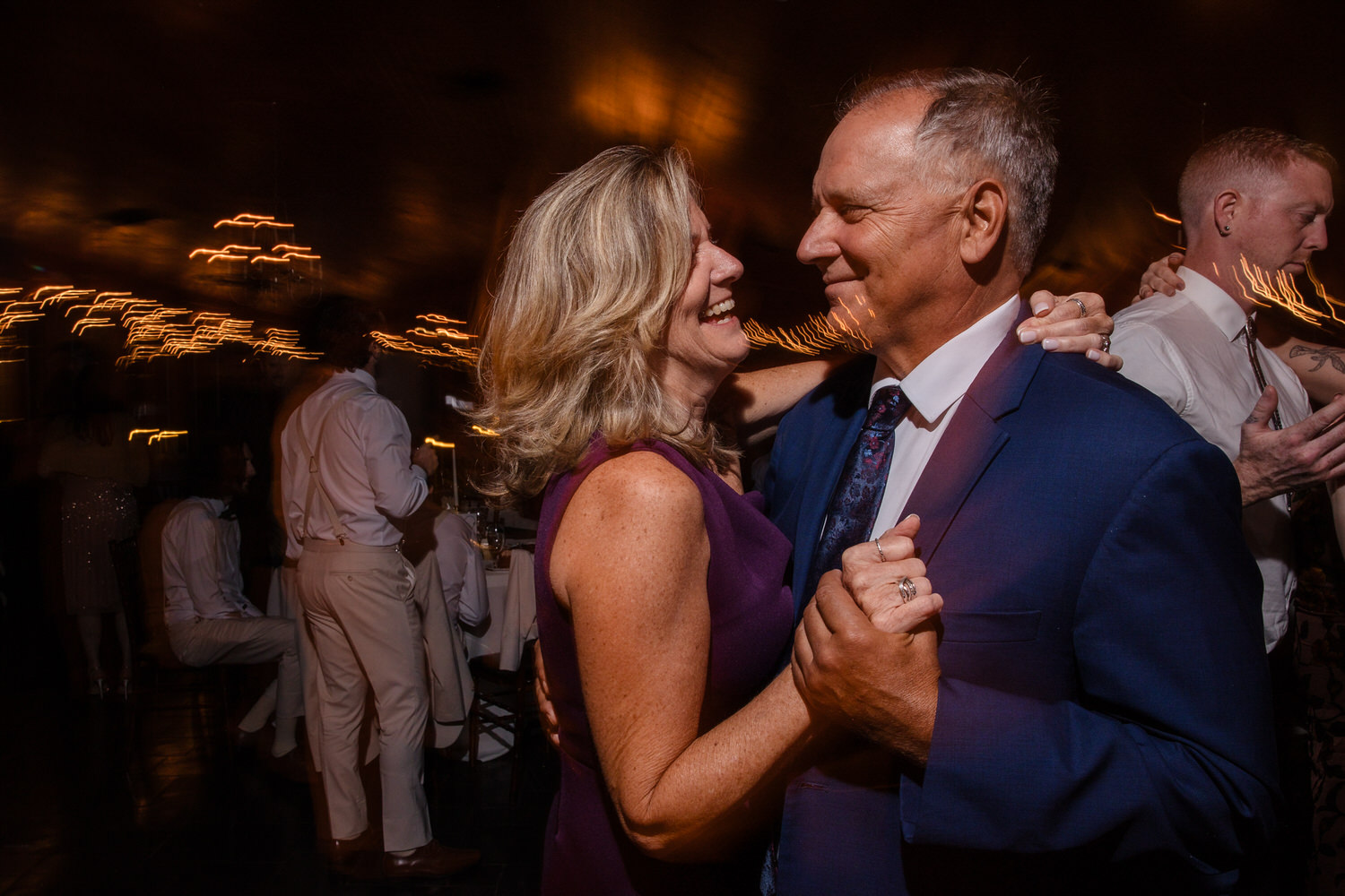 A smiling woman in a purple dress dances with a man in a blue suit at a dimly lit event with blurred light streaks in the background.