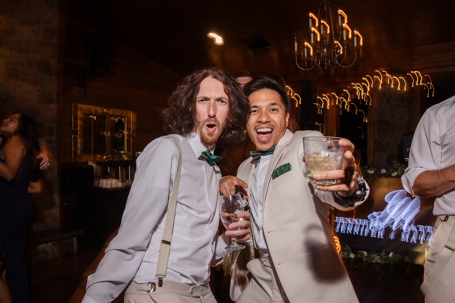 Two men in dress shirts and bow ties toast with glasses at a wedding reception.