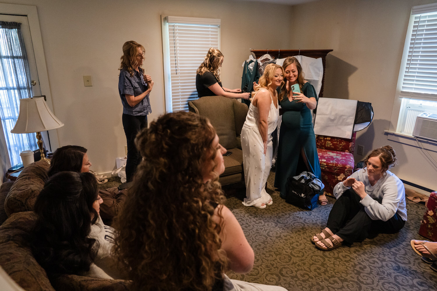 A bride in a white dress and a bridesmaid in a dark green dress take a selfie together in a room filled with wedding party members getting ready.