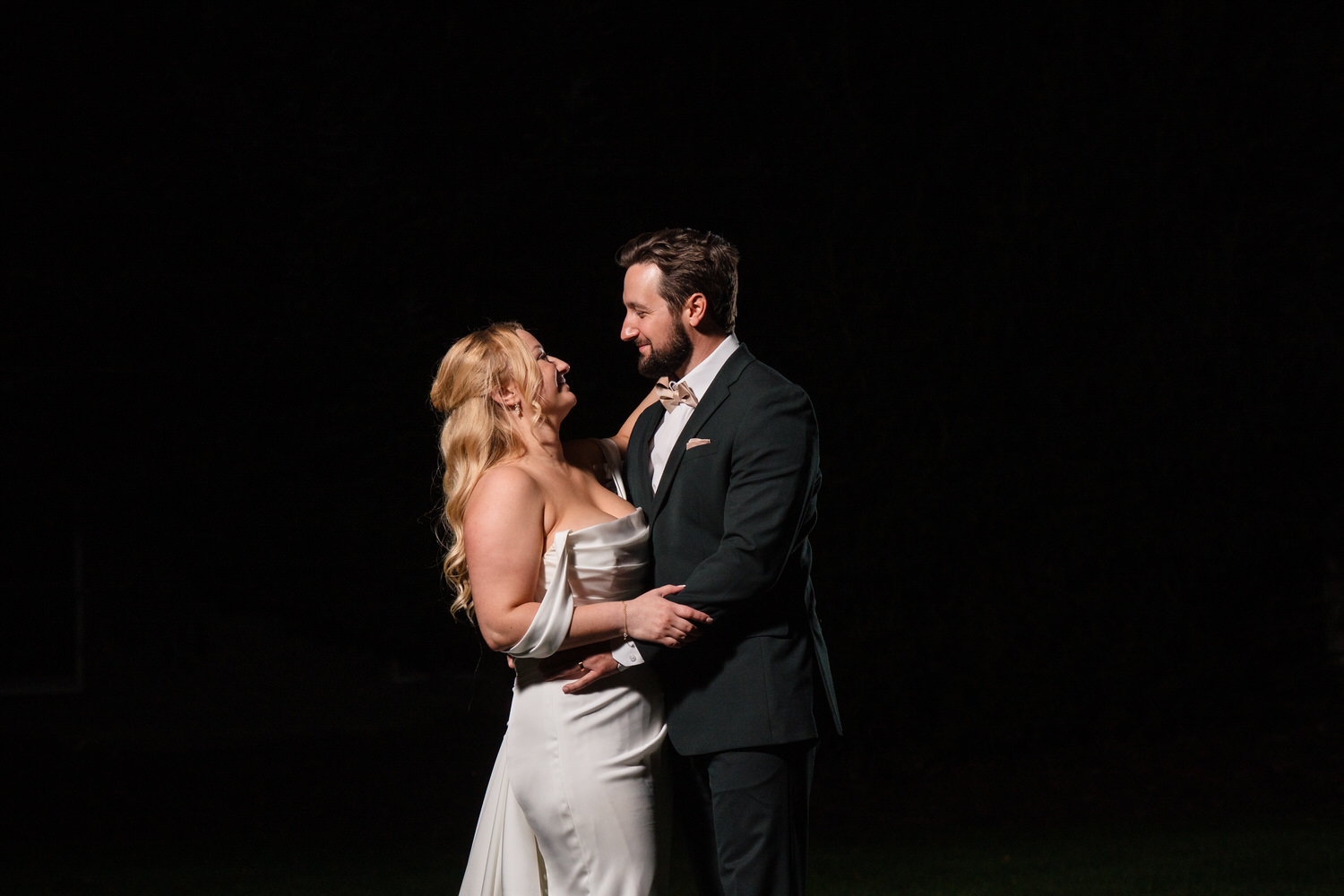 A bride in a white wedding gown and a groom in a dark suit standing face-to-face against a black background.