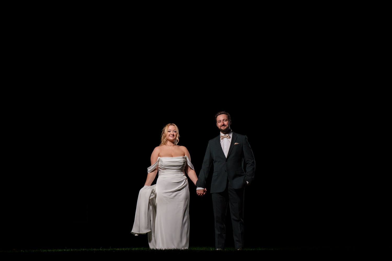 A bride in a white wedding gown and a groom in a dark suit hold hands against a solid black background.