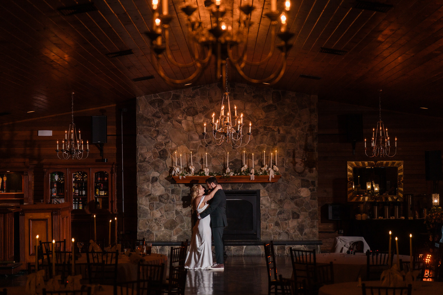 A bride and groom share a slow dance in front of a stone fireplace in a dimly lit, rustic hall decorated with chandeliers.