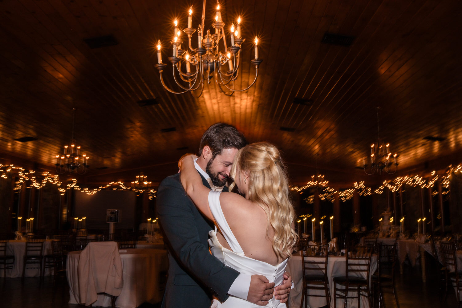 A bride and groom embrace while dancing in a dimly lit banquet hall under hanging chandeliers.